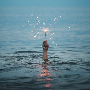 A hand reaches up out of the water, holding a lit sparkler. The rest of the body is submerged. Negative self talk.