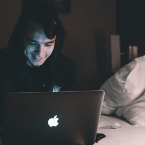 A smilking man uses a laptop while sitting on a bed. Online addiction treatment