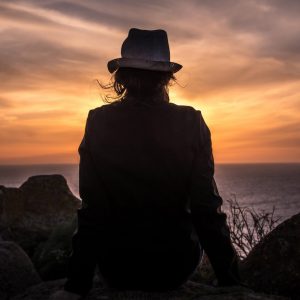 Rear view of a shadowed image. A woman in a hat stares out at the sunset.